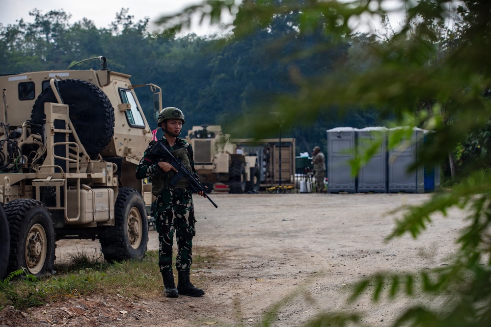 A soldier in green camouflage uniform and helmet stands guard on a dirt road while holding an assault rifle. Behind him are tan/beige military vehicles, likely MRAPs or HMMWVs, along with portable toilets and equipment suggesting a forward operating base or staging area. A second soldier is visible in the background near equipment.