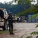 A soldier in green camouflage uniform and helmet stands guard on a dirt road while holding an assault rifle. Behind him are tan/beige military vehicles, likely MRAPs or HMMWVs, along with portable toilets and equipment suggesting a forward operating base or staging area. A second soldier is visible in the background near equipment.