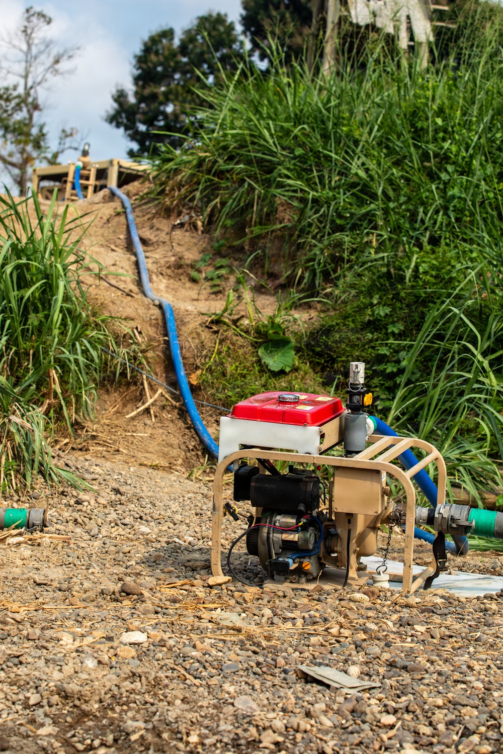 A gasoline-powered water pump with a red fuel tank sits on a gravel surface at the base of an earthen embankment. A blue hose runs from the pump up the slope toward a wooden structure at the top of the hill, suggesting a water transfer or pumping operation. The setting appears to be a rural or semi-rural construction or irrigation site surrounded by dense vegetation.