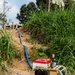 A gasoline-powered water pump with a red fuel tank sits on a gravel surface at the base of an earthen embankment. A blue hose runs from the pump up the slope toward a wooden structure at the top of the hill, suggesting a water transfer or pumping operation. The setting appears to be a rural or semi-rural construction or irrigation site surrounded by dense vegetation.