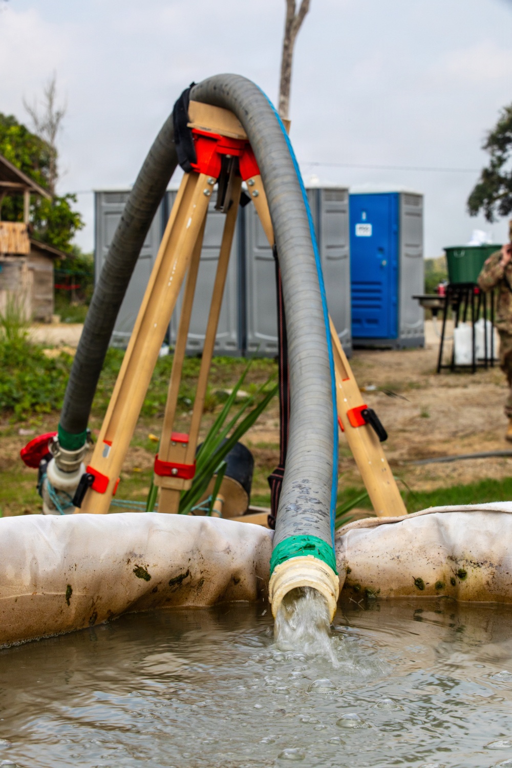 The image shows a water pumping operation in an outdoor field setting, with large flexible hoses mounted on a wooden tripod structure actively discharging water into a fabric-lined containment basin or berm. The setup appears to be a military or humanitarian water supply operation, with portable toilets and temporary infrastructure visible in the background. Water is visibly flowing from the hose outlet into the pooled water below.
