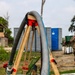 The image shows a water pumping operation in an outdoor field setting, with large flexible hoses mounted on a wooden tripod structure actively discharging water into a fabric-lined containment basin or berm. The setup appears to be a military or humanitarian water supply operation, with portable toilets and temporary infrastructure visible in the background. Water is visibly flowing from the hose outlet into the pooled water below.