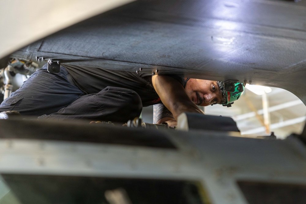 A technician or mechanic is working in a confined space beneath what appears to be a large aircraft or vehicle, lying on their back or side to access components. The person is wearing dark work clothing and has a headlamp or light mounted on their head. The image is shot from a low angle through a narrow gap, creating a dramatic perspective of the maintenance work.