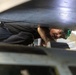 A technician or mechanic is working in a confined space beneath what appears to be a large aircraft or vehicle, lying on their back or side to access components. The person is wearing dark work clothing and has a headlamp or light mounted on their head. The image is shot from a low angle through a narrow gap, creating a dramatic perspective of the maintenance work.