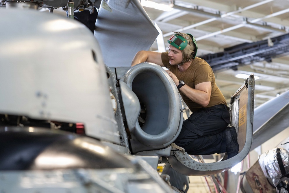 A military aircraft maintenance technician is performing close inspection or maintenance work on what appears to be a jet engine intake or exhaust nozzle of a military aircraft. The work is being conducted inside a hangar facility. The technician is wearing standard naval/military aviation deck crew gear including a cranial helmet with ear protection.