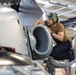 A military aircraft maintenance technician is performing close inspection or maintenance work on what appears to be a jet engine intake or exhaust nozzle of a military aircraft. The work is being conducted inside a hangar facility. The technician is wearing standard naval/military aviation deck crew gear including a cranial helmet with ear protection.