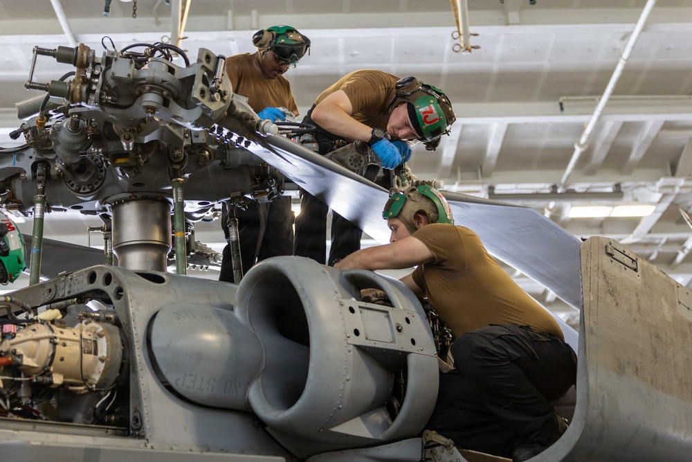 Three military aviation maintenance technicians are performing maintenance work on a military helicopter, likely in a hangar bay aboard a naval vessel or air station. The personnel are wearing standard-issue brown t-shirts and green cranial helmets with ear protection, consistent with U.S. Navy or Marine Corps aviation maintenance crews. They are working on the rotor head assembly and engine/transmission area of what appears to be a military helicopter.