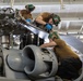 Three military aviation maintenance technicians are performing maintenance work on a military helicopter, likely in a hangar bay aboard a naval vessel or air station. The personnel are wearing standard-issue brown t-shirts and green cranial helmets with ear protection, consistent with U.S. Navy or Marine Corps aviation maintenance crews. They are working on the rotor head assembly and engine/transmission area of what appears to be a military helicopter.