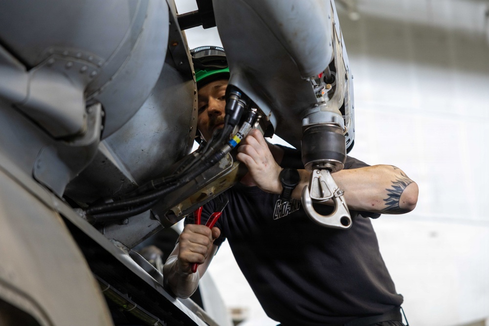 A male technician or mechanic is performing maintenance work on what appears to be a military or commercial aircraft, working inside a tight access panel or engine compartment area. He is using tools including pliers and what appears to be a pneumatic or power tool while closely inspecting internal components. The work is being conducted in an indoor hangar environment.