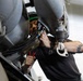 A male technician or mechanic is performing maintenance work on what appears to be a military or commercial aircraft, working inside a tight access panel or engine compartment area. He is using tools including pliers and what appears to be a pneumatic or power tool while closely inspecting internal components. The work is being conducted in an indoor hangar environment.