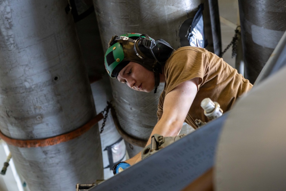 A young male technician or military personnel is working in a confined mechanical space, likely aboard a naval vessel or aircraft. He is wearing a green helmet with ear protection and a tan/brown t-shirt while performing maintenance or inspection work. The environment contains large cylindrical pipes, chains, and metal structural components.