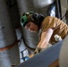 A young male technician or military personnel is working in a confined mechanical space, likely aboard a naval vessel or aircraft. He is wearing a green helmet with ear protection and a tan/brown t-shirt while performing maintenance or inspection work. The environment contains large cylindrical pipes, chains, and metal structural components.