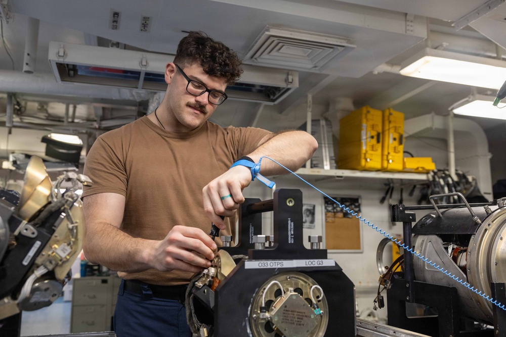 A young male technician wearing a brown t-shirt and glasses is performing maintenance or repair work on a piece of mechanical or electronic equipment in what appears to be a naval vessel workspace. He is wearing an anti-static wrist strap (blue ESD strap) on his left wrist, indicating he is working with sensitive electronic components. The workspace contains various technical equipment, yellow storage cases, and mechanical components consistent with a military ship's interior.