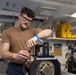 A young male technician wearing a brown t-shirt and glasses is performing maintenance or repair work on a piece of mechanical or electronic equipment in what appears to be a naval vessel workspace. He is wearing an anti-static wrist strap (blue ESD strap) on his left wrist, indicating he is working with sensitive electronic components. The workspace contains various technical equipment, yellow storage cases, and mechanical components consistent with a military ship's interior.