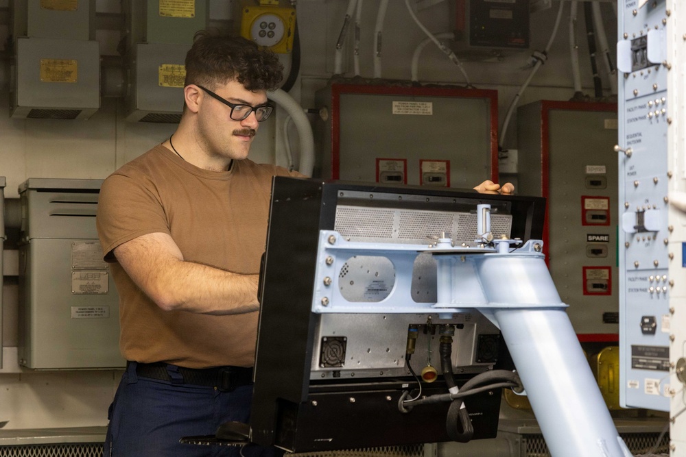A young male technician wearing glasses, a brown t-shirt, and dark pants is working on an electronic equipment unit mounted on a pedestal or stand. The setting appears to be a military or industrial facility, likely aboard a naval vessel, based on the visible electrical panels, conduit, and red-bordered control boxes in the background. He appears to be inspecting or servicing the rear/interior of the equipment.