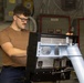 A young male technician wearing glasses, a brown t-shirt, and dark pants is working on an electronic equipment unit mounted on a pedestal or stand. The setting appears to be a military or industrial facility, likely aboard a naval vessel, based on the visible electrical panels, conduit, and red-bordered control boxes in the background. He appears to be inspecting or servicing the rear/interior of the equipment.