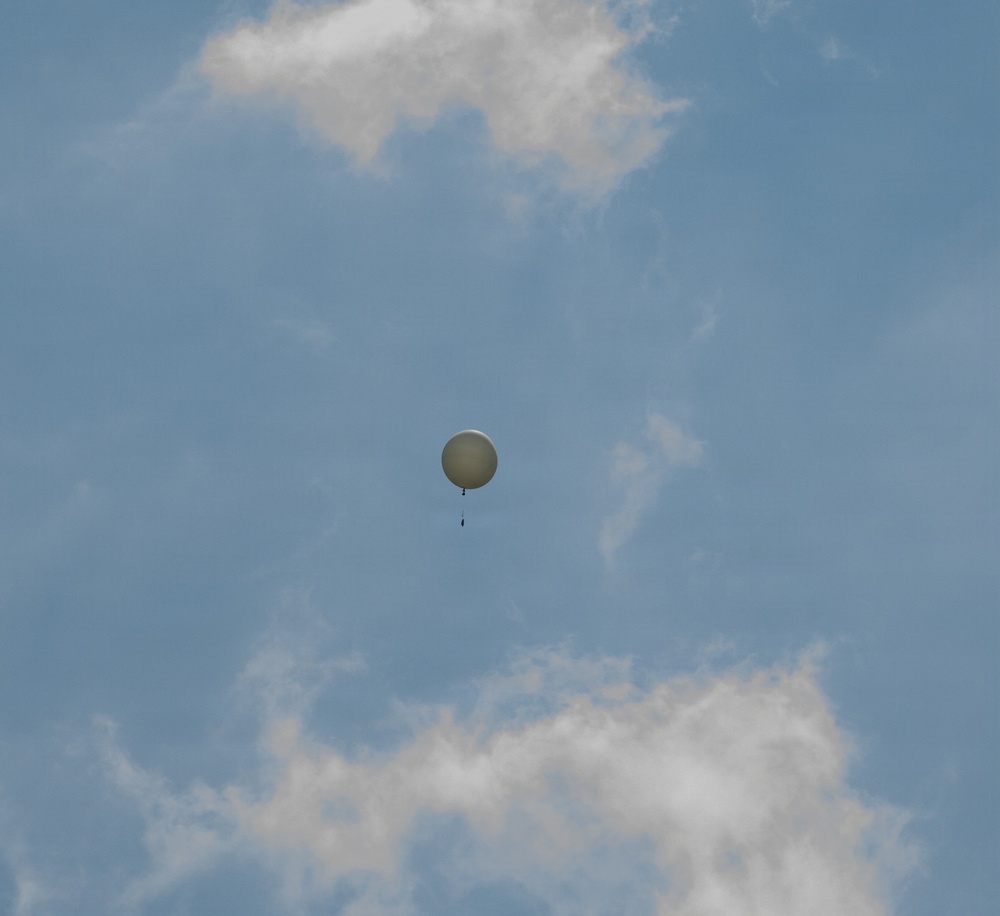 A single white/light-colored spherical balloon is visible ascending through a partly cloudy blue sky. A small payload or instrument package appears to be suspended beneath the balloon via a thin line or cord. The image appears to depict a weather or radiosonde balloon used for atmospheric data collection.