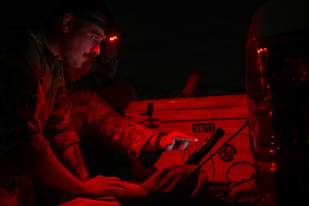A military personnel member is working at night under red light illumination, using a tablet device while wearing a headlamp with a red light mode. The scene appears to be a field operation or exercise, with equipment including a YETI cooler visible in the background. Red lighting is used to preserve night vision during tactical operations.