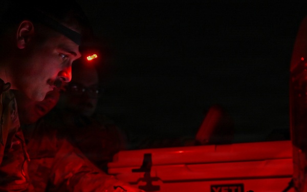 A military personnel member is working at night under red light illumination, using a tablet device while wearing a headlamp with a red light mode. The scene appears to be a field operation or exercise, with equipment including a YETI cooler visible in the background. Red lighting is used to preserve night vision during tactical operations.
