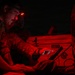 A military personnel member is working at night under red light illumination, using a tablet device while wearing a headlamp with a red light mode. The scene appears to be a field operation or exercise, with equipment including a YETI cooler visible in the background. Red lighting is used to preserve night vision during tactical operations.