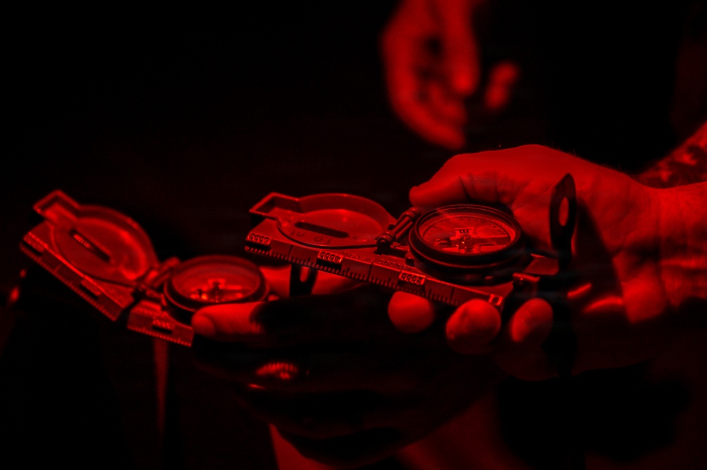 A person is holding two military-style lensatic compasses under red light illumination against a dark background. The red lighting is consistent with night navigation practices, where red light preserves night vision while still allowing instrument reading. The compasses appear to be open and in use for land navigation purposes.