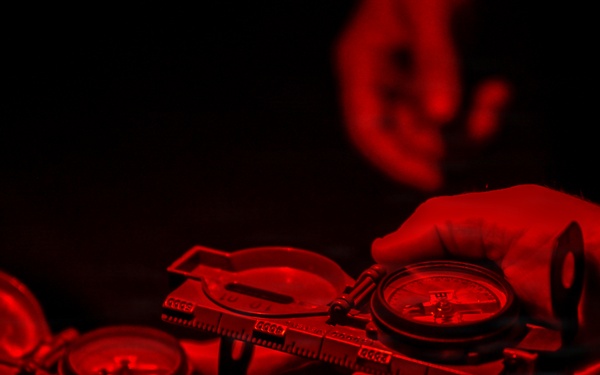 A person is holding two military-style lensatic compasses under red light illumination against a dark background. The red lighting is consistent with night navigation practices, where red light preserves night vision while still allowing instrument reading. The compasses appear to be open and in use for land navigation purposes.