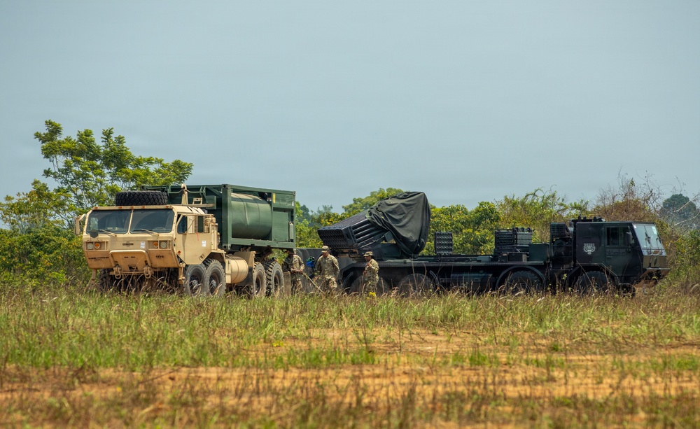 The image shows a military field operation involving two large military trucks in an open grassy area with tree line in the background. A tan/beige HEMTT (Heavy Expanded Mobility Tactical Truck) is positioned alongside a dark-colored heavy transport vehicle carrying what appears to be a covered large military system on a flatbed. Several soldiers in military uniforms are visible between the vehicles, apparently conducting equipment transfer or setup operations.