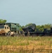 The image shows a military field operation involving two large military trucks in an open grassy area with tree line in the background. A tan/beige HEMTT (Heavy Expanded Mobility Tactical Truck) is positioned alongside a dark-colored heavy transport vehicle carrying what appears to be a covered large military system on a flatbed. Several soldiers in military uniforms are visible between the vehicles, apparently conducting equipment transfer or setup operations.