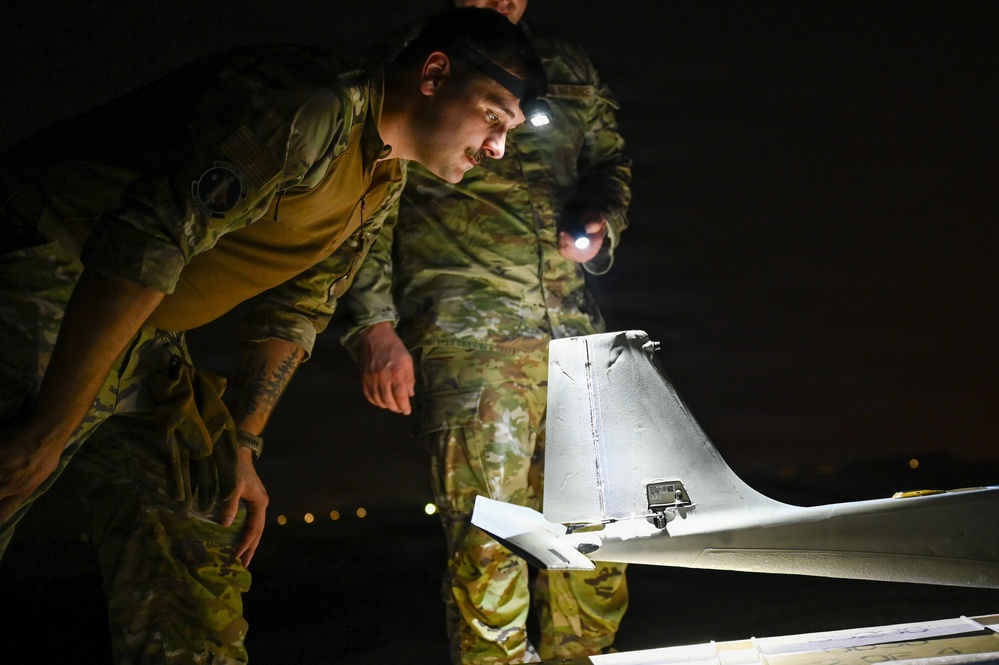 Two military personnel in camouflage uniforms are conducting a nighttime inspection of a small unmanned aerial vehicle (UAV/drone). One soldier is leaning in closely to examine the aircraft while another illuminates the area using a handheld flashlight and a headlamp. The setting appears to be an outdoor tarmac or field environment in complete darkness.