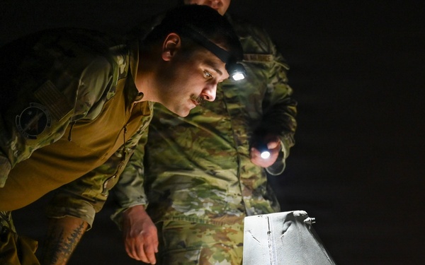 Two military personnel in camouflage uniforms are conducting a nighttime inspection of a small unmanned aerial vehicle (UAV/drone). One soldier is leaning in closely to examine the aircraft while another illuminates the area using a handheld flashlight and a headlamp. The setting appears to be an outdoor tarmac or field environment in complete darkness.