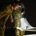 Two military personnel in camouflage uniforms are conducting a nighttime inspection of a small unmanned aerial vehicle (UAV/drone). One soldier is leaning in closely to examine the aircraft while another illuminates the area using a handheld flashlight and a headlamp. The setting appears to be an outdoor tarmac or field environment in complete darkness.
