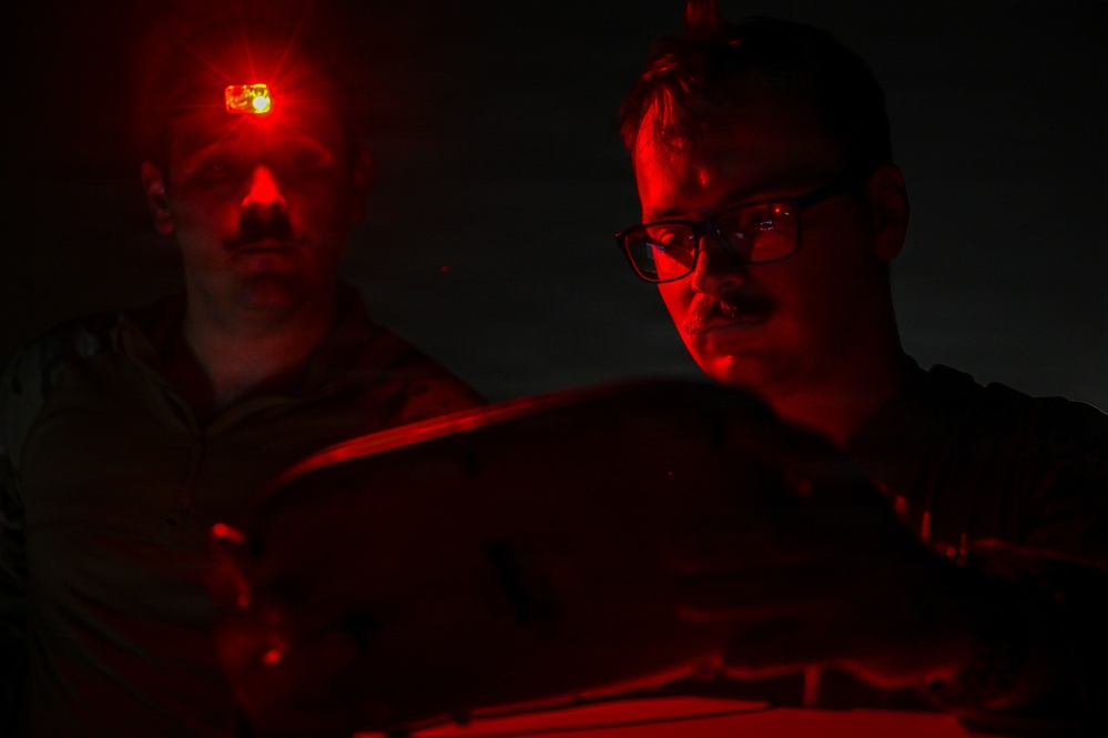 Two individuals are photographed in near-total darkness, illuminated primarily by red light. The person on the left has a small red LED light mounted on their head, while the person on the right wears glasses and appears to be focused on something below the frame. The dramatic red lighting and dark environment suggest a nighttime operational or tactical setting.