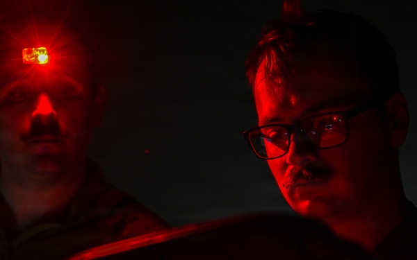 Two individuals are photographed in near-total darkness, illuminated primarily by red light. The person on the left has a small red LED light mounted on their head, while the person on the right wears glasses and appears to be focused on something below the frame. The dramatic red lighting and dark environment suggest a nighttime operational or tactical setting.