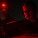 Two individuals are photographed in near-total darkness, illuminated primarily by red light. The person on the left has a small red LED light mounted on their head, while the person on the right wears glasses and appears to be focused on something below the frame. The dramatic red lighting and dark environment suggest a nighttime operational or tactical setting.