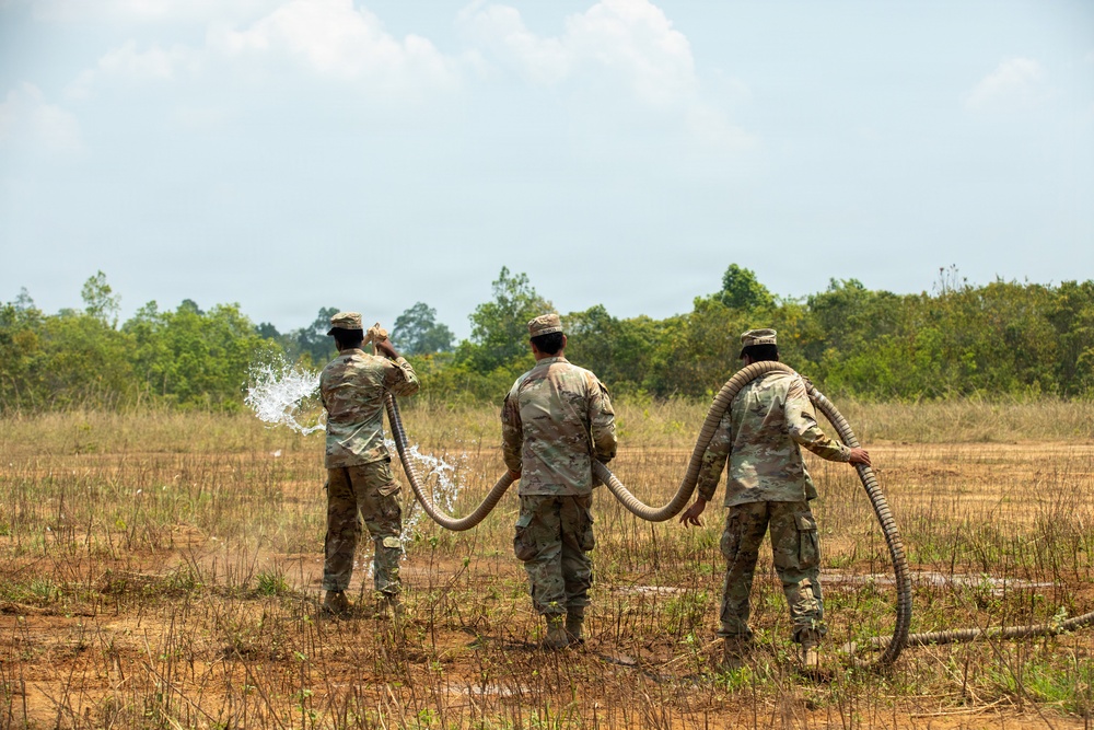 Three military personnel in camouflage uniforms are handling large flexible hoses in an open field, with water visibly spraying from the hose held by the leftmost individual. They appear to be conducting a water distribution or field engineering exercise in a rural, semi-arid outdoor environment. The soldiers are viewed from behind, standing in a line across a dry, partially vegetated field.