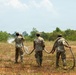 Three military personnel in camouflage uniforms are handling large flexible hoses in an open field, with water visibly spraying from the hose held by the leftmost individual. They appear to be conducting a water distribution or field engineering exercise in a rural, semi-arid outdoor environment. The soldiers are viewed from behind, standing in a line across a dry, partially vegetated field.