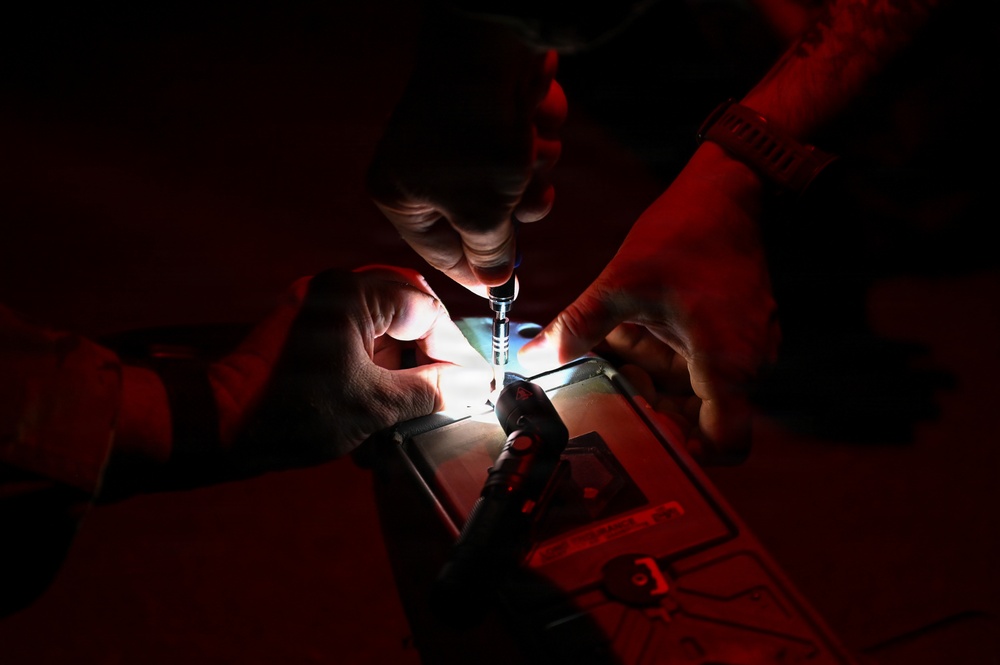 Two individuals are working together in a very dark environment, illuminated primarily by a small focused light source from below. One person is holding what appears to be a syringe or precision instrument over a lit surface or device. The scene suggests a delicate technical or medical procedure being performed under low-light conditions.