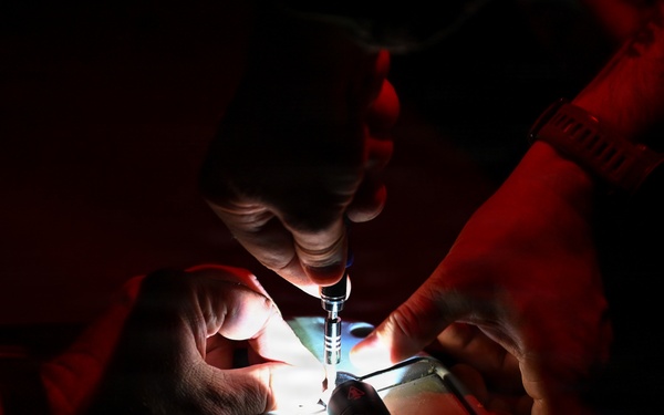 Two individuals are working together in a very dark environment, illuminated primarily by a small focused light source from below. One person is holding what appears to be a syringe or precision instrument over a lit surface or device. The scene suggests a delicate technical or medical procedure being performed under low-light conditions.