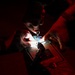 Two individuals are working together in a very dark environment, illuminated primarily by a small focused light source from below. One person is holding what appears to be a syringe or precision instrument over a lit surface or device. The scene suggests a delicate technical or medical procedure being performed under low-light conditions.