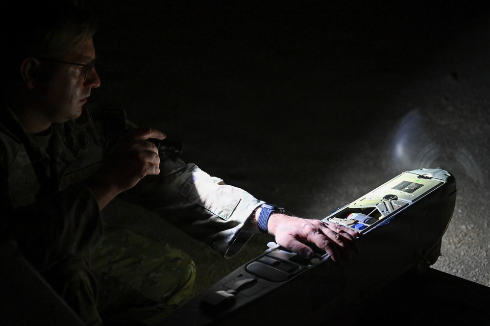 A person in military camouflage uniform is working with what appears to be a small fixed-wing drone or UAV in low-light conditions. They are illuminating the device with a flashlight or torch while appearing to inspect or configure it. The scene suggests a nighttime military or training operation involving unmanned aerial system preparation.