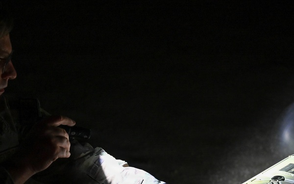 A person in military camouflage uniform is working with what appears to be a small fixed-wing drone or UAV in low-light conditions. They are illuminating the device with a flashlight or torch while appearing to inspect or configure it. The scene suggests a nighttime military or training operation involving unmanned aerial system preparation.