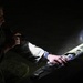 A person in military camouflage uniform is working with what appears to be a small fixed-wing drone or UAV in low-light conditions. They are illuminating the device with a flashlight or torch while appearing to inspect or configure it. The scene suggests a nighttime military or training operation involving unmanned aerial system preparation.