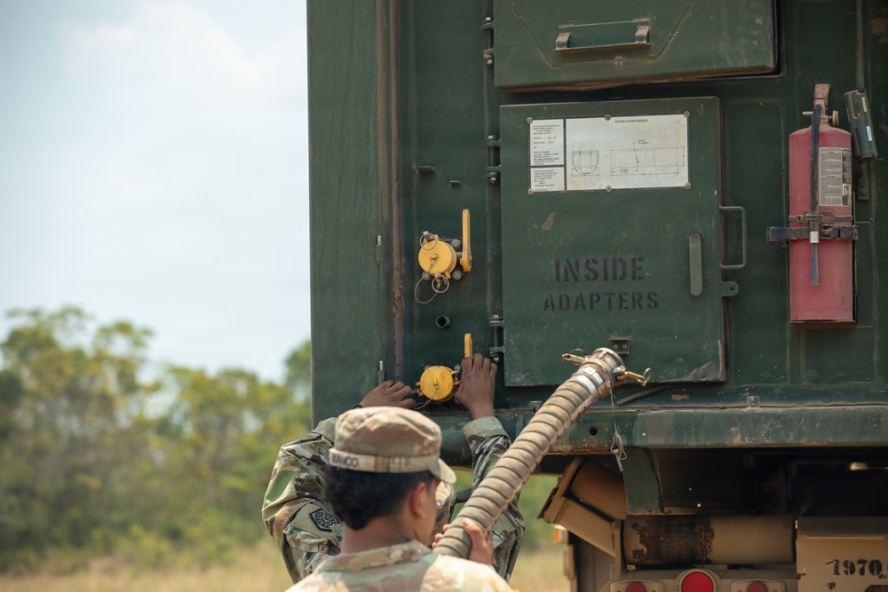 A military service member in camouflage uniform is working at the rear of a large green military vehicle or container. The soldier appears to be connecting or manipulating a hose/coupling at a panel labeled 'INSIDE ADAPTERS.' A red fire extinguisher is mounted on the exterior of the vehicle, and yellow valve/coupling components are visible on the panel.