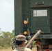 A military service member in camouflage uniform is working at the rear of a large green military vehicle or container. The soldier appears to be connecting or manipulating a hose/coupling at a panel labeled 'INSIDE ADAPTERS.' A red fire extinguisher is mounted on the exterior of the vehicle, and yellow valve/coupling components are visible on the panel.