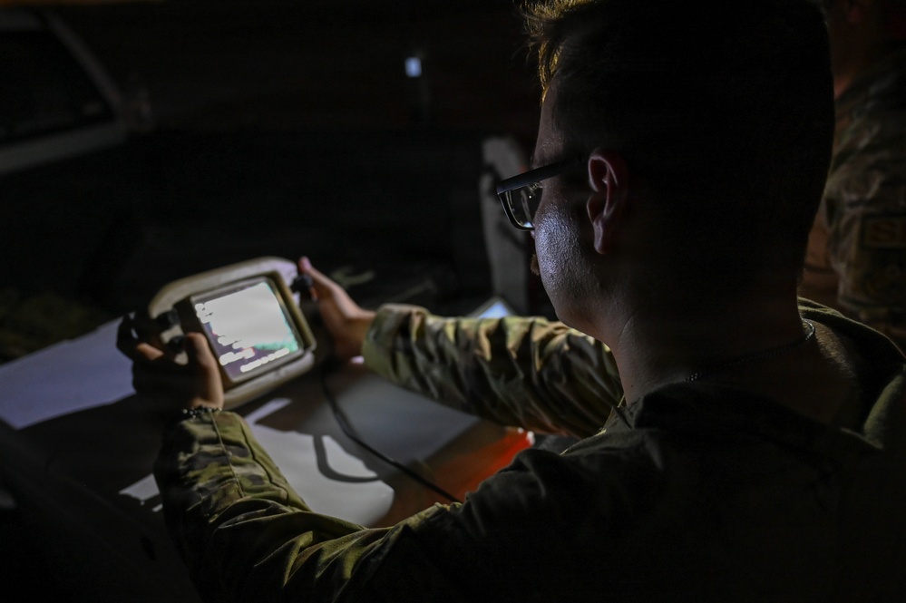 A person in military camouflage uniform is operating a ruggedized handheld device with a small illuminated screen in a low-light environment. The individual is wearing glasses and appears focused on the device's display. The setting suggests a tactical or field operations context.