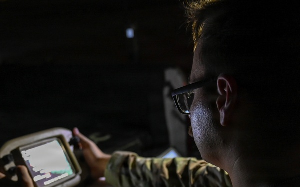 A person in military camouflage uniform is operating a ruggedized handheld device with a small illuminated screen in a low-light environment. The individual is wearing glasses and appears focused on the device's display. The setting suggests a tactical or field operations context.