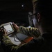 A person in military camouflage uniform is operating a ruggedized handheld device with a small illuminated screen in a low-light environment. The individual is wearing glasses and appears focused on the device's display. The setting suggests a tactical or field operations context.
