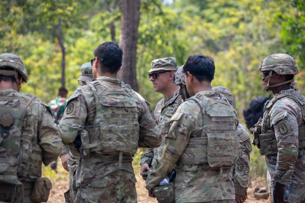 A group of U.S. Army soldiers in MultiCam/OCP camouflage uniforms and body armor are gathered in a wooded outdoor area, appearing to be in a briefing or discussion. The central figure is wearing a patrol cap and sunglasses with a visible U.S. Army name tape, while others wear helmets. The setting and mixed group composition suggest a joint training exercise or field operation.