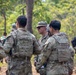 A group of U.S. Army soldiers in MultiCam/OCP camouflage uniforms and body armor are gathered in a wooded outdoor area, appearing to be in a briefing or discussion. The central figure is wearing a patrol cap and sunglasses with a visible U.S. Army name tape, while others wear helmets. The setting and mixed group composition suggest a joint training exercise or field operation.