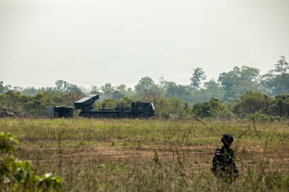 The image shows a military field exercise or deployment in a tropical or subtropical open terrain environment. A large truck-mounted multiple launch rocket system (MLRS) is positioned in the middle distance, with a soldier in camouflage uniform visible in the foreground. The setting appears to be a military training ground or operational area bordered by dense treeline.